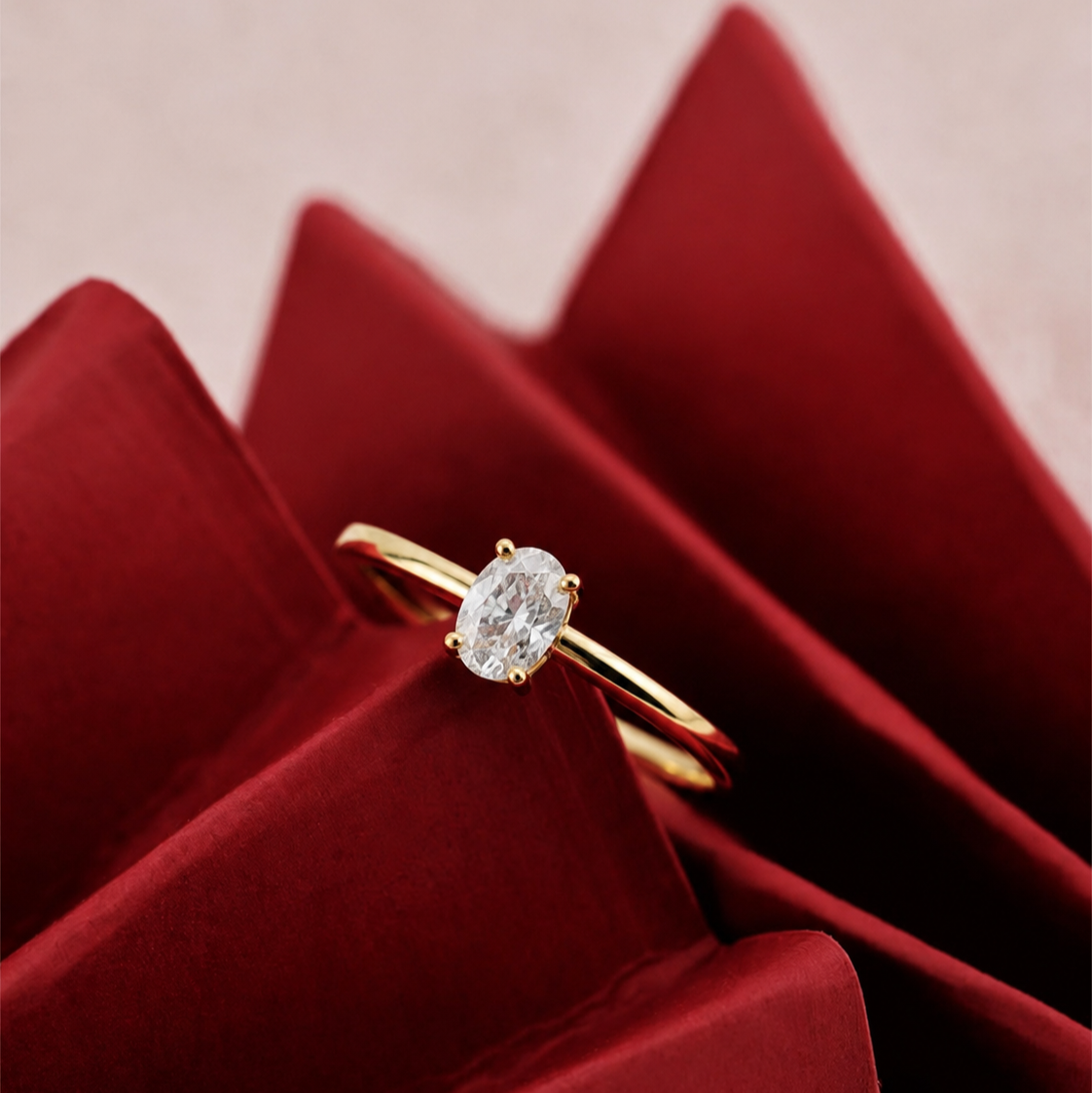 Gold ring with an oval diamond solitaire resting on deep red satin fabric, photographed in a luxury close-up style.
