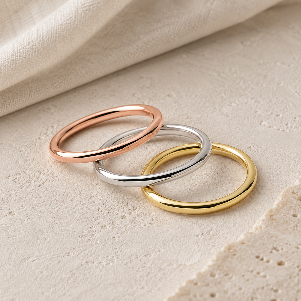 Three polished minimalist rings in rose gold, silver, and yellow gold, arranged together on a textured cream stone surface with soft linen fabric in the background.