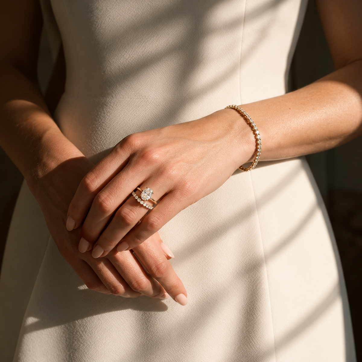 A close-up image of a woman standing in an ivory dress with her hands gently crossed. She is wearing a diamond engagement ring paired with a slim diamond wedding band, along with a delicate diamond bracelet, all shown in warm natural light.