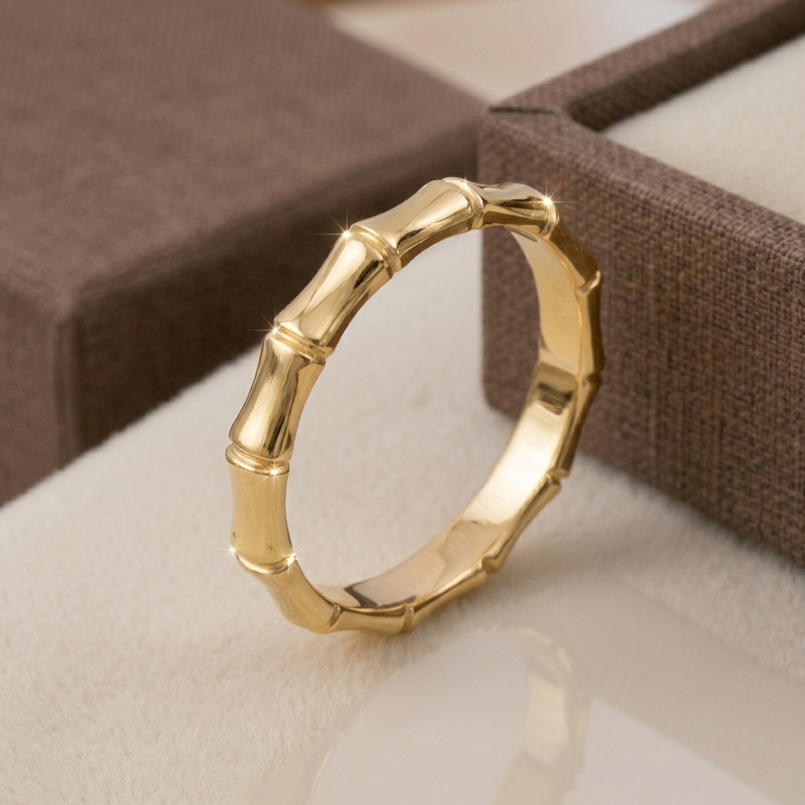 A unique yellow gold bamboo-inspired stackable fashion ring. The segmented band, designed to resemble a bamboo stalk with polished nodes, rests on a plush cream surface next to an open brown textured jewelry box, casting a reflection.
