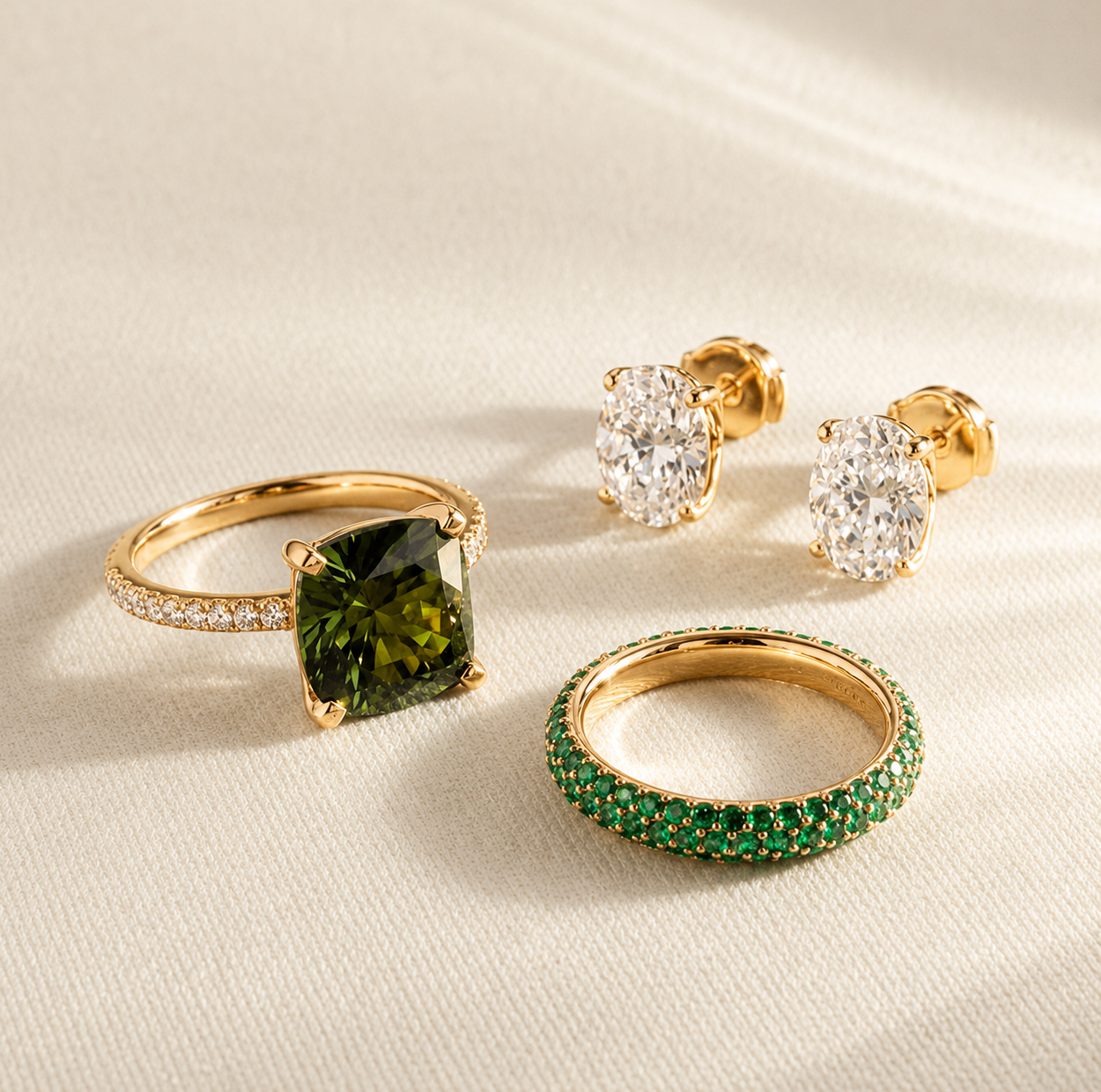 Gold jewelry set with green gemstone rings and oval diamond earrings arranged on a light cream fabric background with soft natural shadows.