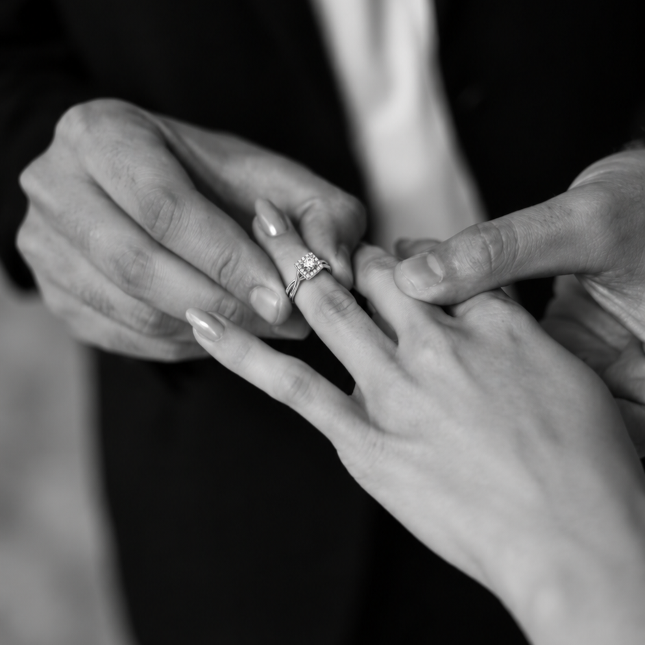 Black-and-white close-up of a couple’s hands as a diamond engagement ring is gently placed on the ring finger.
