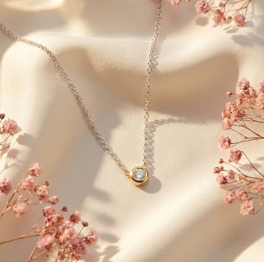 Delicate gold diamond pendant necklace on soft ivory satin, surrounded by pink dried flowers in warm natural light.