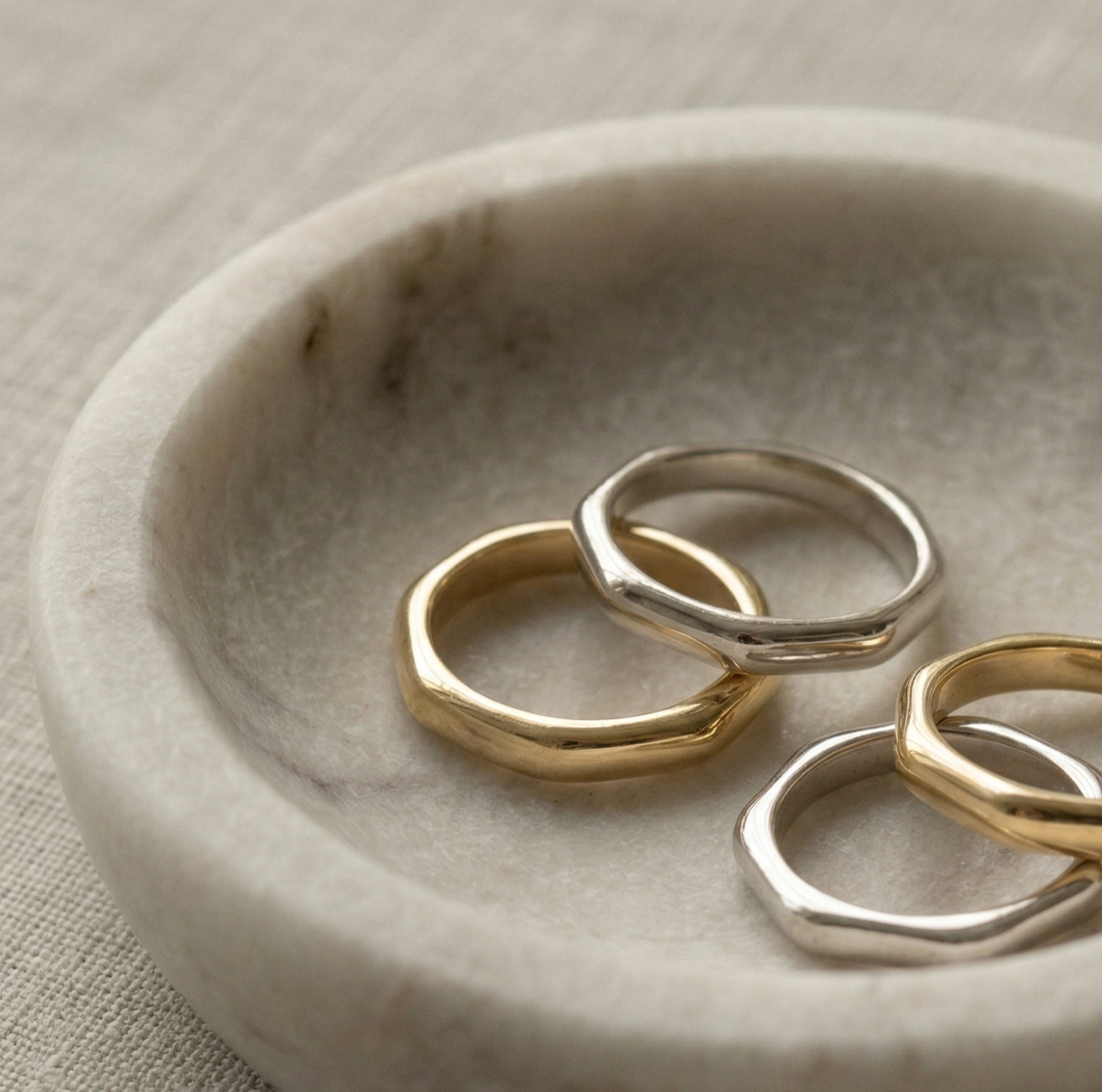 A luxury close-up of four minimalist 18k gold bands—two yellow gold and two white gold - displayed in a textured marble bowl.